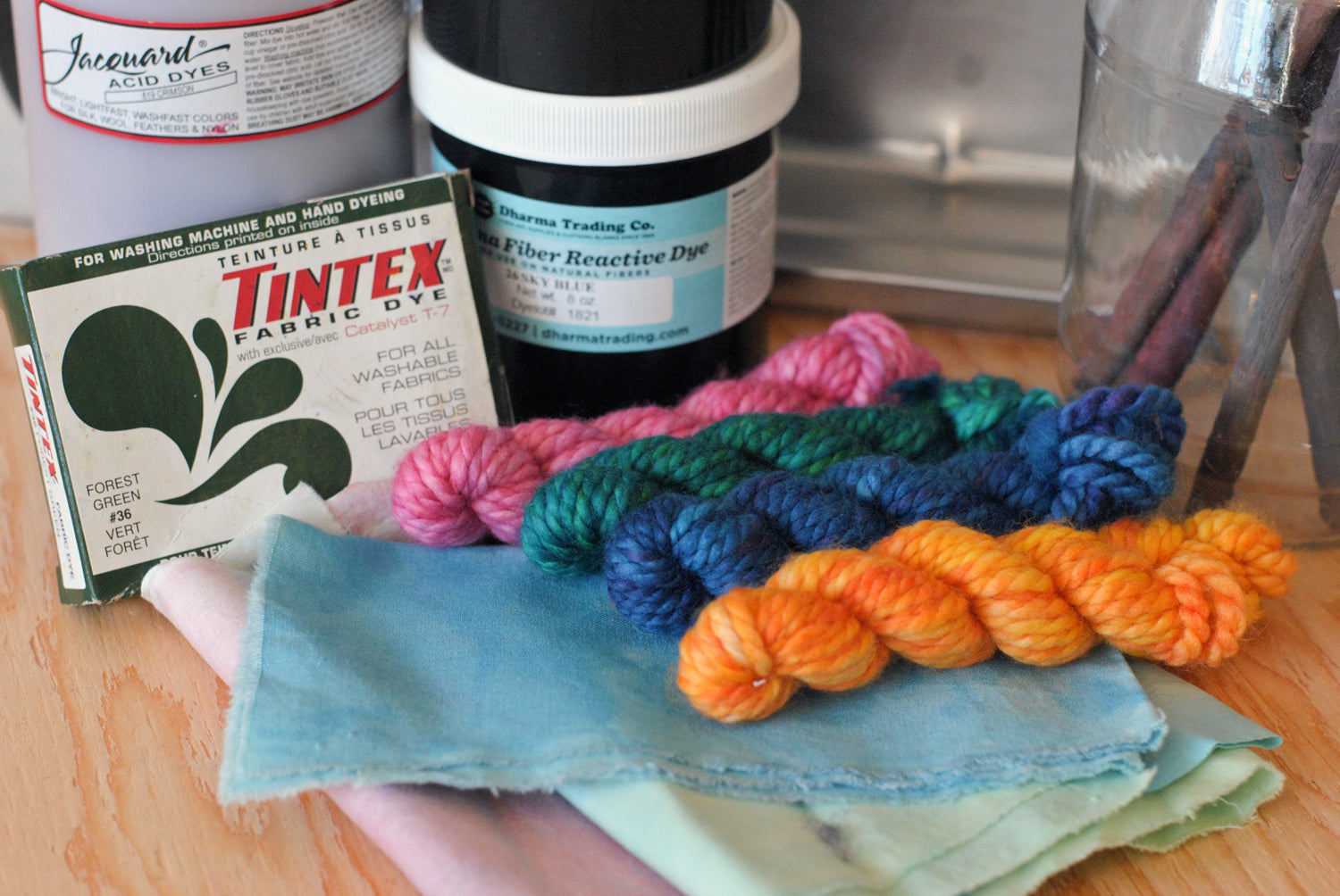 Four mini-skeins of yarn hand-dyed in different colours resting on top of hand-dyed fabric swatches with various types of dye in the background