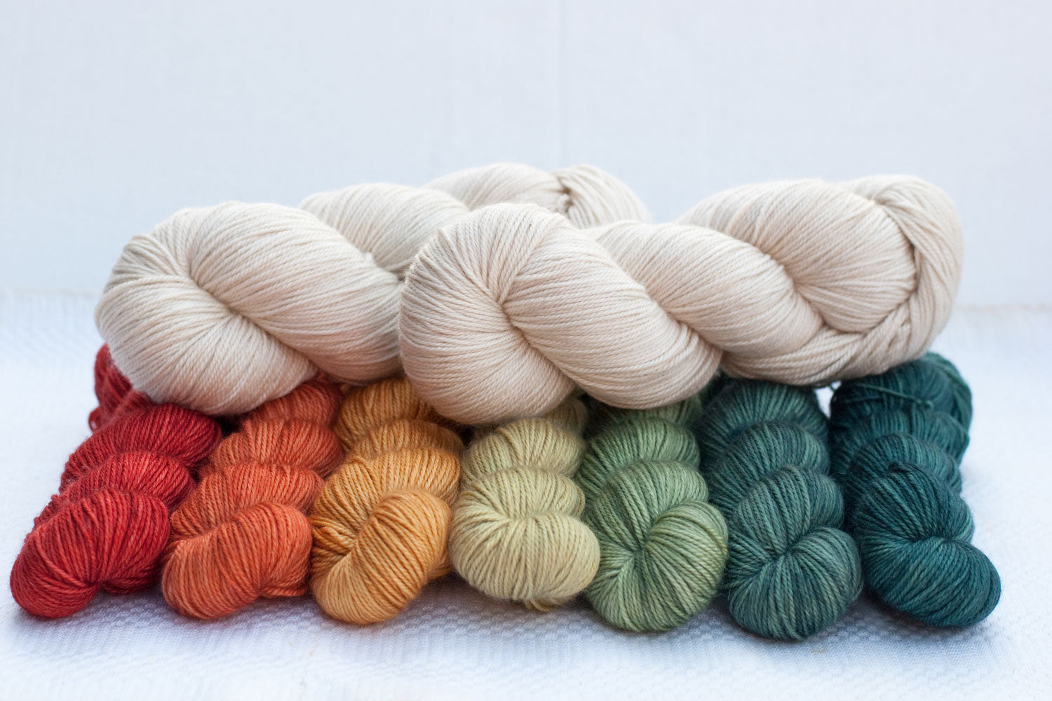 two full skeins of cream yarn arranged on top of seven gradient skeins from red-orange through yellow to green