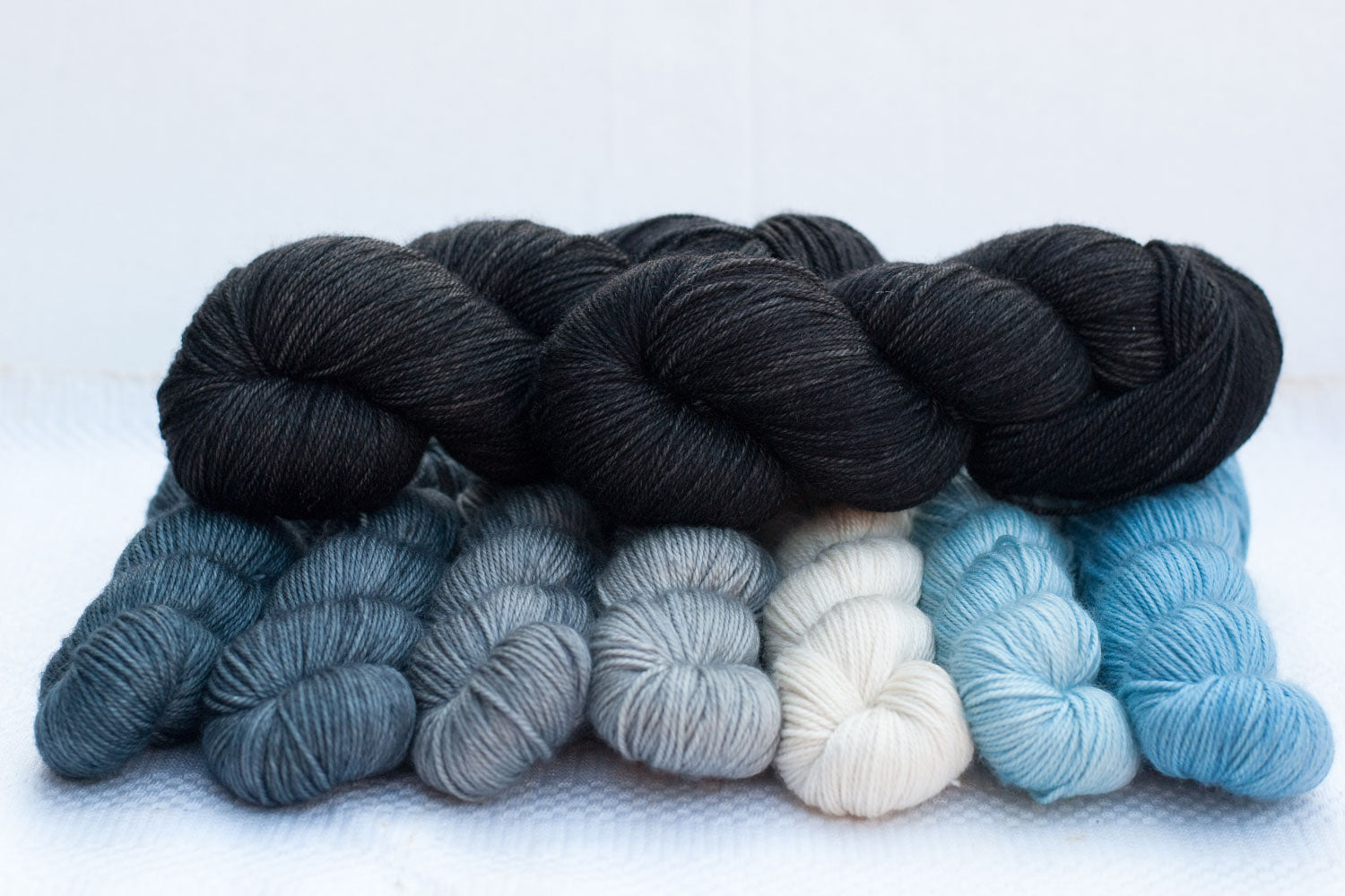 2 full skeins of black hand-dyed yarn arranged on top of grey, white and blue gradient set